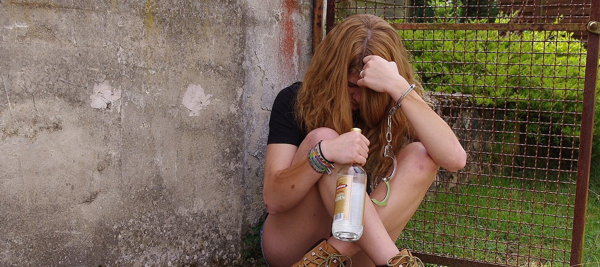 Eine Jugendliche sitzt mit einer Flasche Korn und Handschellen auf dem Boden
