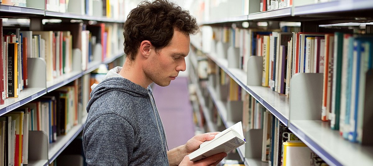 In der Uni-Bibliothek steht ein junger Mann zwischen den Regalen und liest ein Buch