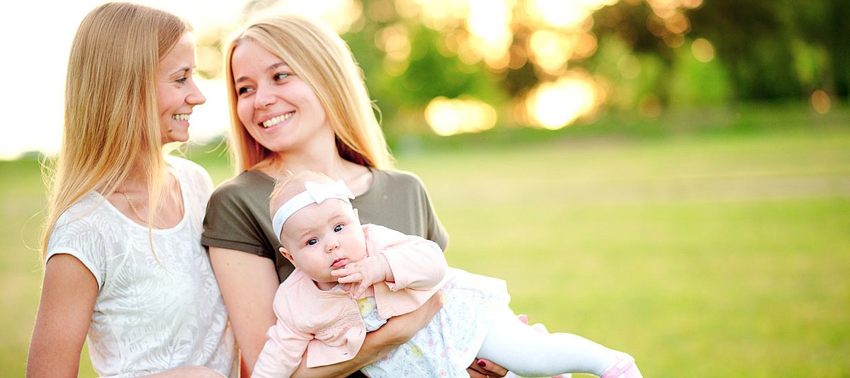 Zwei lesbische Frauen tragen lachend ein Baby im Park.