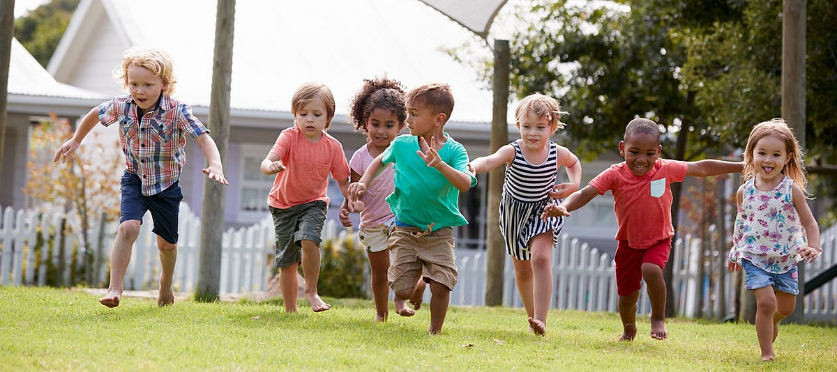 Eine Gruppe Kinder unterschiedlicher Herkunft im Kindergartenalter läuft fröhlich über eine Weise im Garten. 
