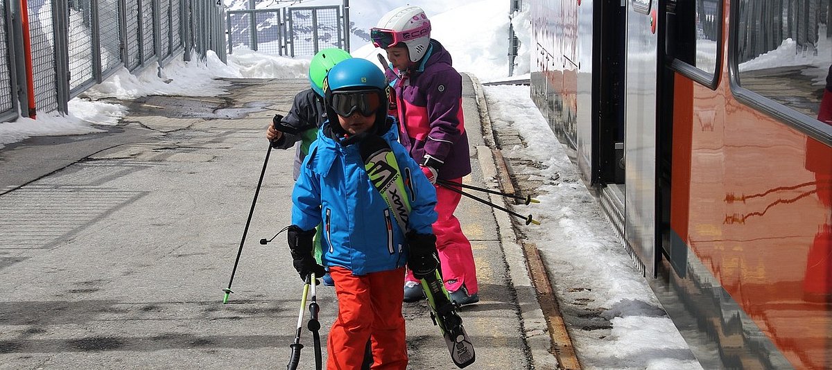 Drei Kinder in voller Skiausrüstung sind in einem verschneiten Bergbahnhof aus einer roten Bahn gestiegen.