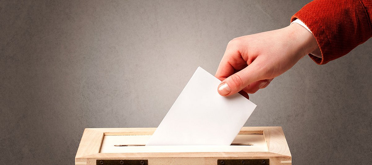Eine Hand wirft einen Wahlzettel in eine Wahlurne. 