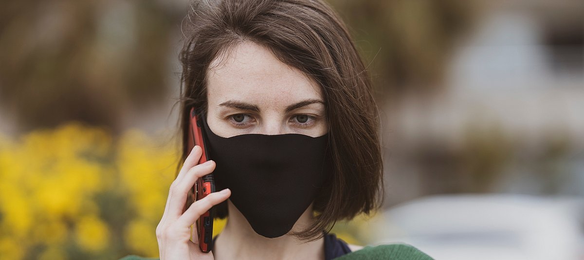 Eine junge Frau mit schwarzer Schutzmaske aus Stoff telefoniert mit ihrem Handy