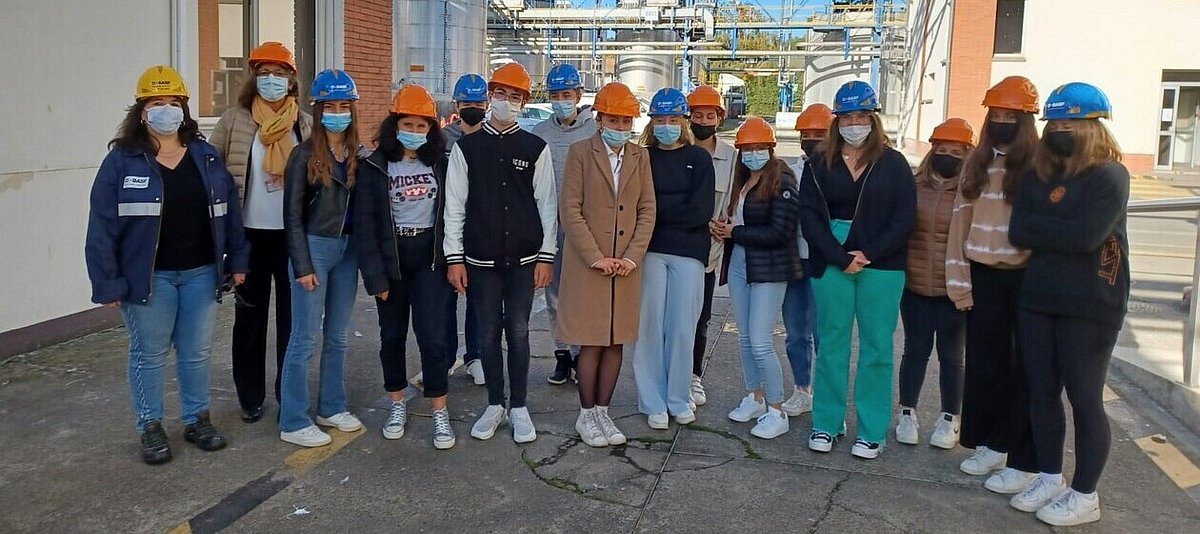 Eine Gruppe Jugendlicher steht mit Atemschutzmasken und Sicherheitshelmen auf dem herbstlich kalten Werksgelände einer Chemiefabrik.
