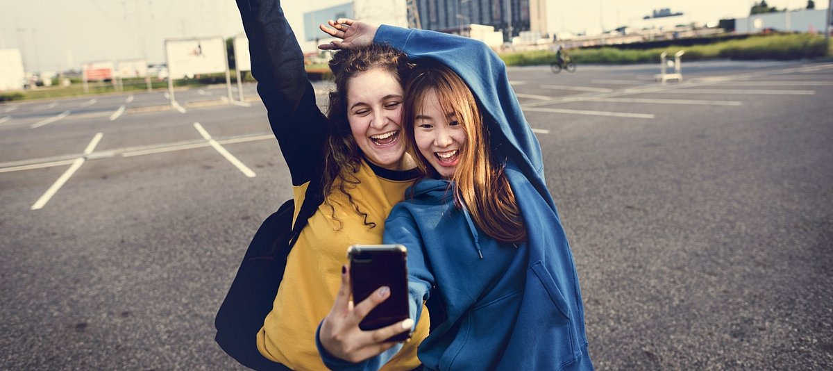 Zwei Jugendliche auf einem verlassenen Parkplatz lachen ausgelassen und machen ein gemeinsames Selfie