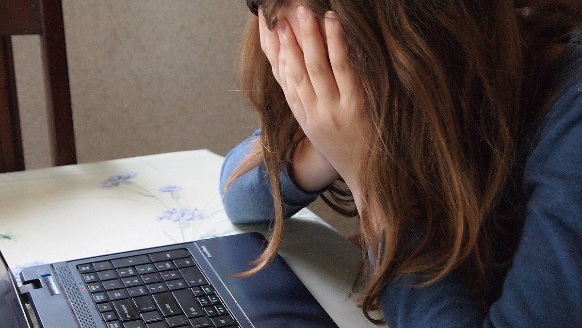 Eine Jugendliche sitzt vor dem Laptop und hält mit geneigtem Kopf verzweifelt die Hände vors Gesicht