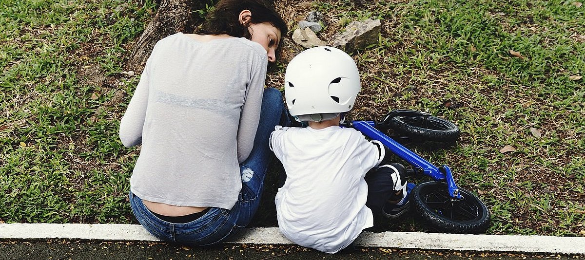 Ein Kind mit Sturzhelm und Laufrad sitzt auf der Bordsteinkante, daneben sitzt eine Erwachsene und spricht mit dem Kind