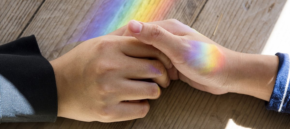 Zwei Hände halten einander auf einer Tischplatte, es sind Regenbogenfarben zu sehen