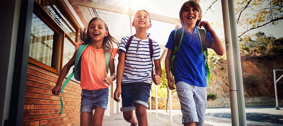Drei Kinder kommen mit ihren Rucksäcke lachend aus einem Schulgebäude