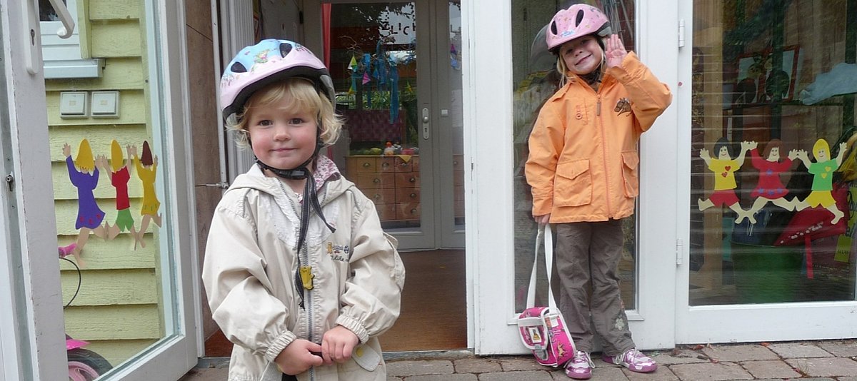 Zwei Kindergartenkinder stehen mit Fahrradhelmen und Taschen vor ihrer Einrichtung