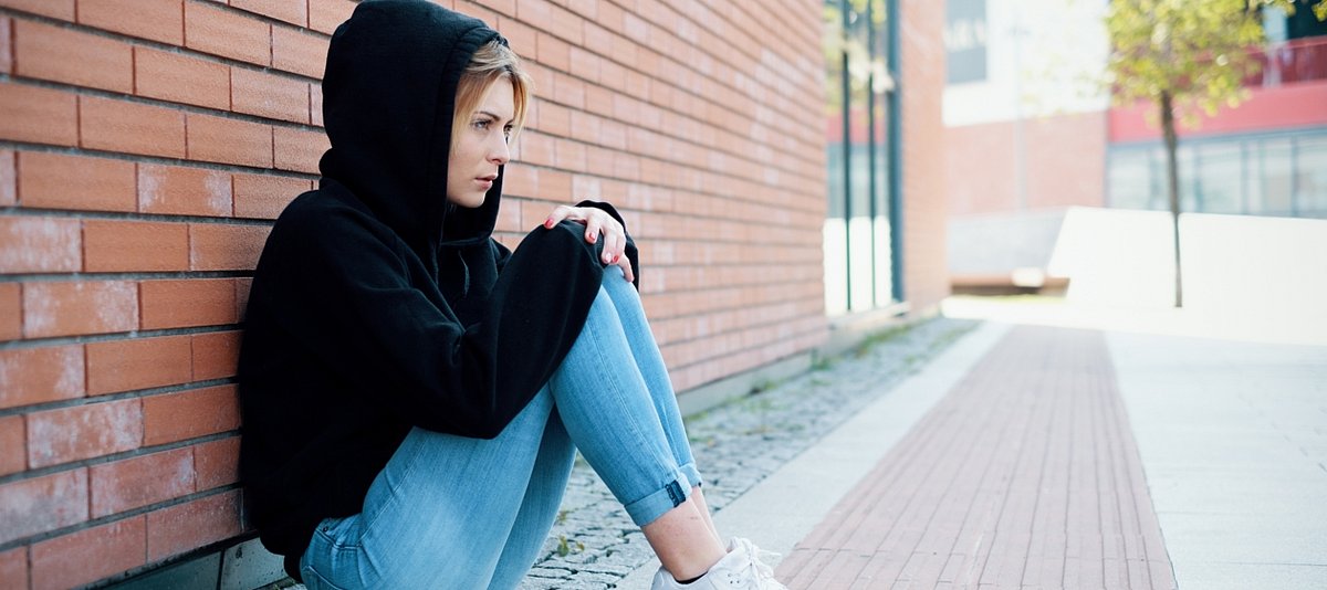Eine Jugendliche in Jeans und Kapuzenpulli gekleidet sitzt an einer Hauswand un d schaut ernst nach vorne