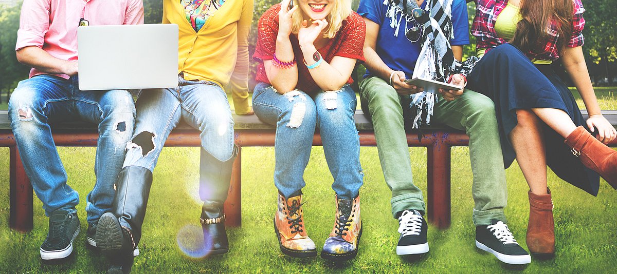 Bunt gekleidete Jugendliche sitzen nebeneinander auf einer Bank, man sieht nur die Beine