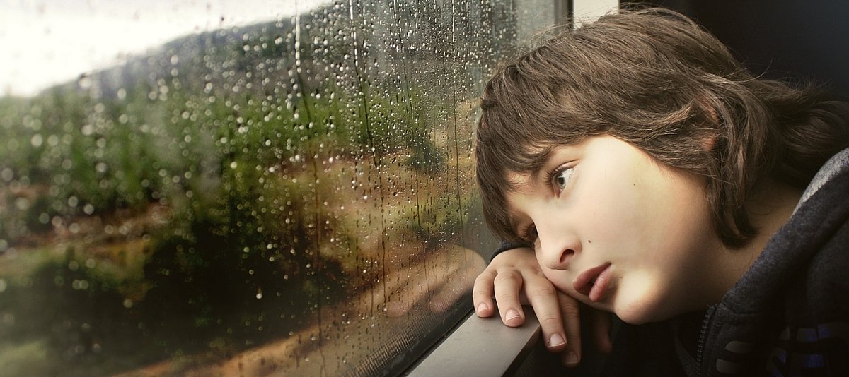 Ein Junge schaut ernst und nachdenklich aus einem Zugfenster, auf dem REgentropfen zu sehen sind