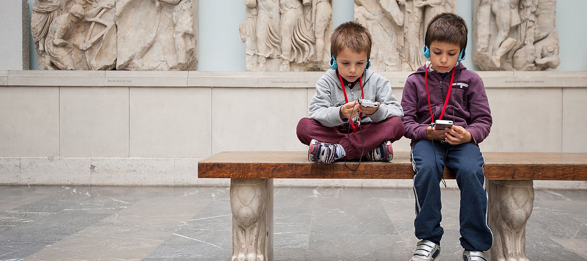 Zwei Jungen sitzen im Museum auf einer Bank und hören dem Audioguide zu.