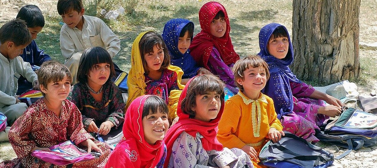 Afghanische Schulkinder sitzen mit bunter Kleidung und Schulutensilien auf dem Boden