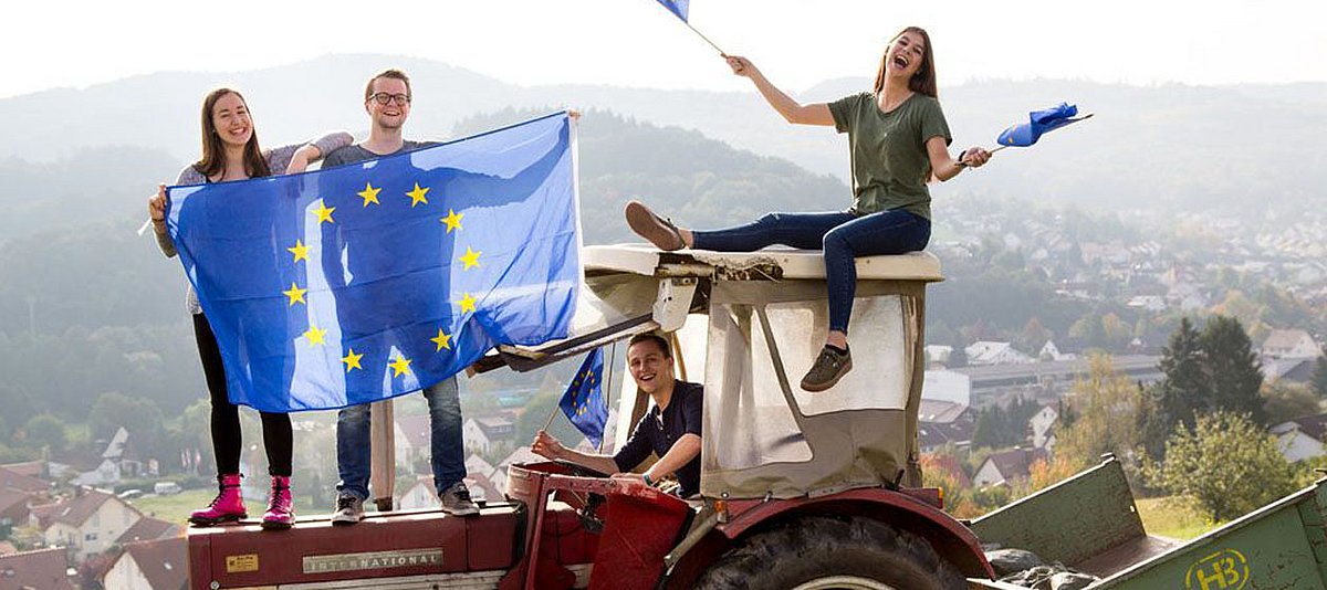 Jugendliche sitzen und stehen lachend auf einem Traktor und wehen mit Europafahnen. 