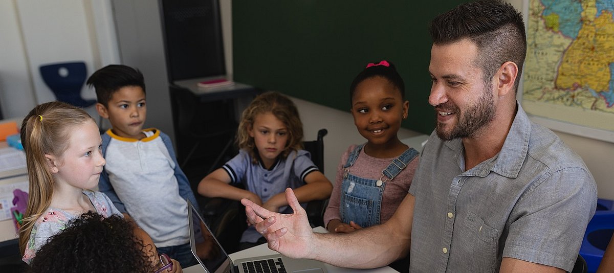 Ein Erwachsener sitzt an einem Laptop und erklärt etwas, um ihn herum sind Kinder, die aufmerksam zuhören