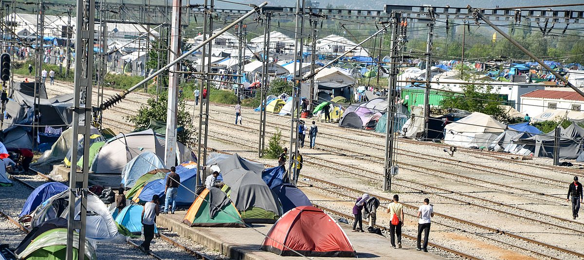Zelte mit Menschen unterschiedlicher Herkunft stehen auf Schienen mit einem Lager im Hintergrund. 