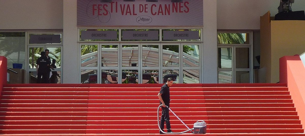 Ein Mitarbeiter in Schwarz reinigt mit einem Staubsauger den roten Teppich auf den Stufen vor dem Festivalgebäude in Cannes.