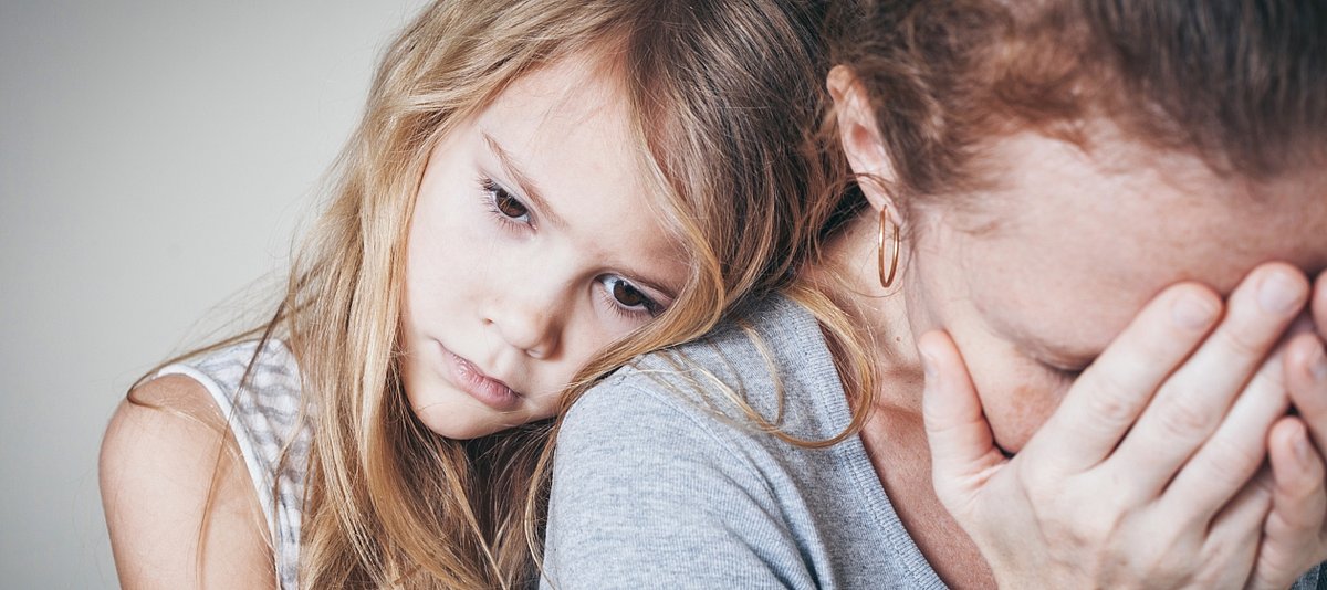 Ein trauriges Mädchen umarmt von hinten ihre Mutter, die verzweifelt ihre Hände vors Gesicht hält