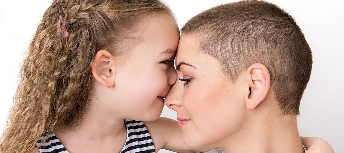 Eine Mutter mit kurzen Haaren wie nach einer Chemotherapie wird von ihrer kleinenTochter umarmt und auf die Nase geküsst.