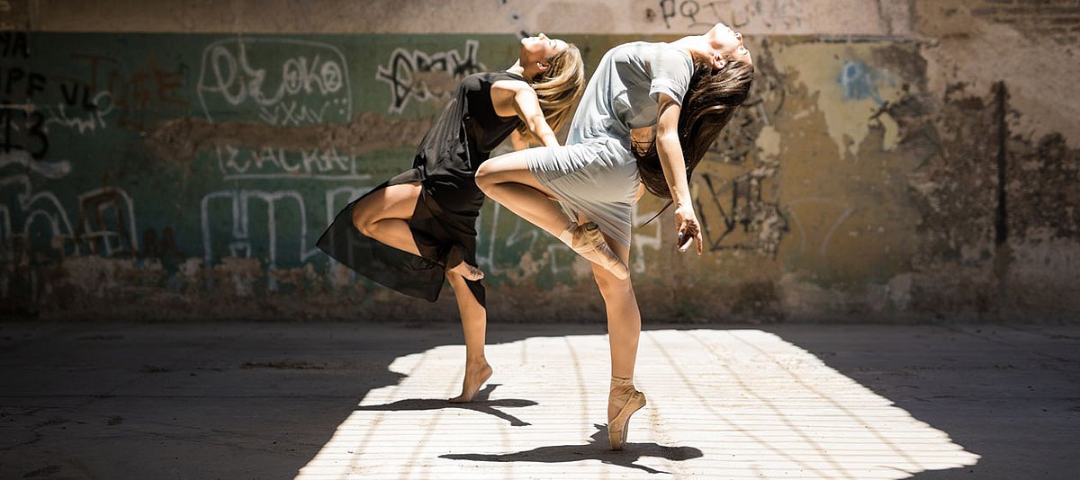 Zwei Junge Frauen tanzen synchron vor einer Mauer mit Graffiti.