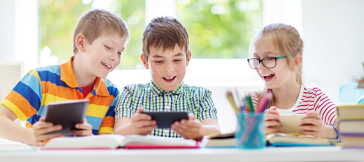 Drei Kinder sitzen in der Schule an einem Tisch mit Büchern und Stiften und schauen sich lachend auf Tablets etwas an