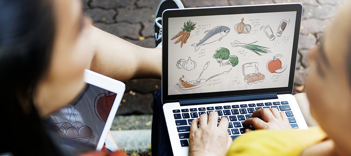 Zwei junge Menschen sitzen mit Laptop und Tablet draußen und diskutieren, Auf dem Bildschirm ist Obst und Gemüse zu erkennen