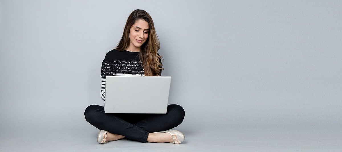 Eine junge Frau sitzt im Schneidersitz und liest etwas auf ihrem Laptop, der auf ihren Beinen steht