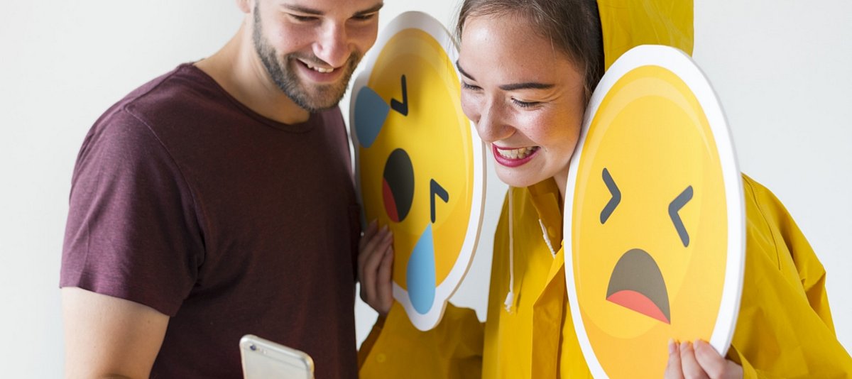 Mann und Frau halten Emoji-Schilder und schauen lachend auf ein Smartphone