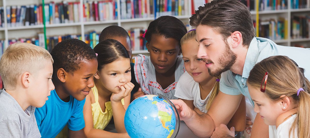 Schüler/-innen schauen gemeinsam mit ihrem Lehrer in einer Bibliothek auf einen Globus. 