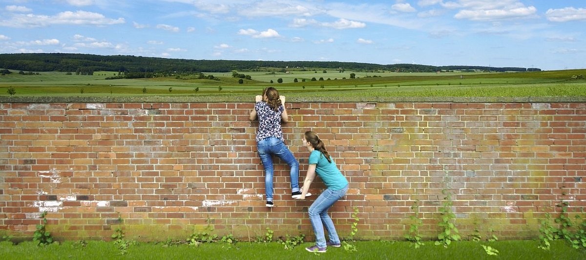 Ein Mädchen schaut mithilfe einer Räuberleiter über eine Mauer auf ein ländliches Gebiet
