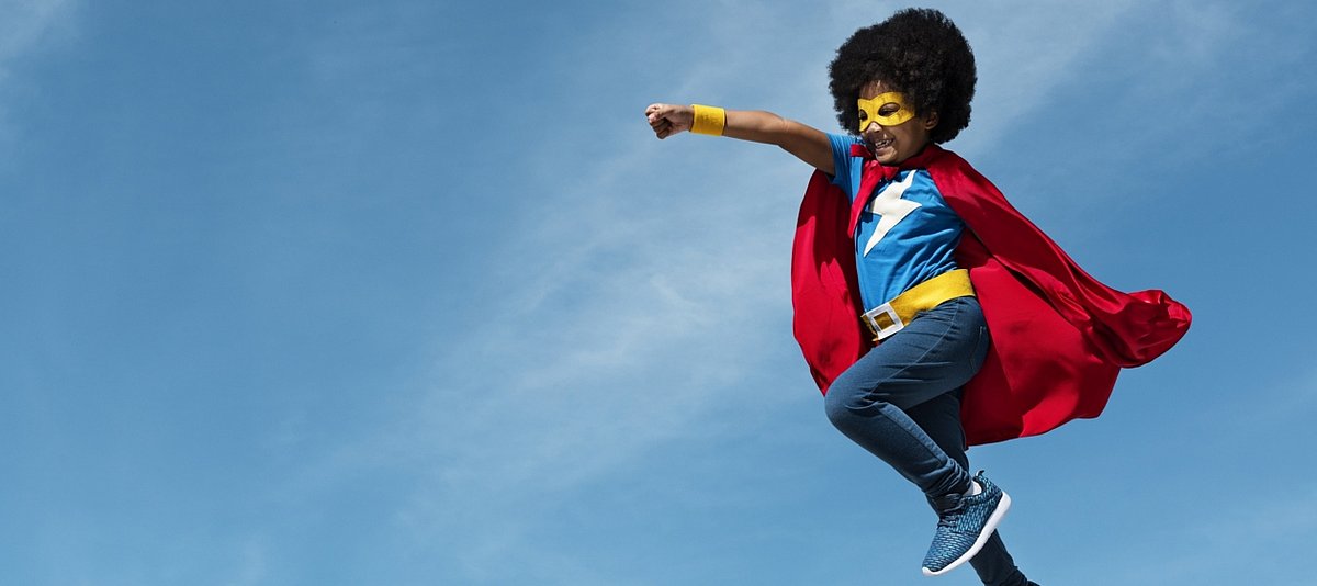 Ein als Superheld verkleideter dunkelhäutiger Junge springt kraftvoll in die Luft