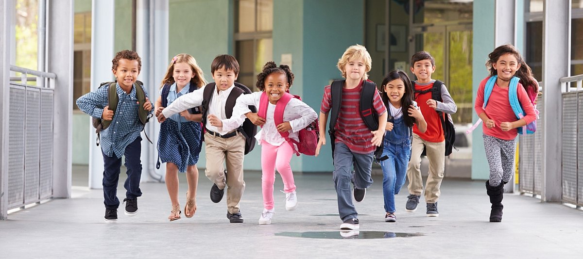 Eine Gruppe Grundschulkinder mit Schulranzen rennt lachend aus dem Schulgebäude