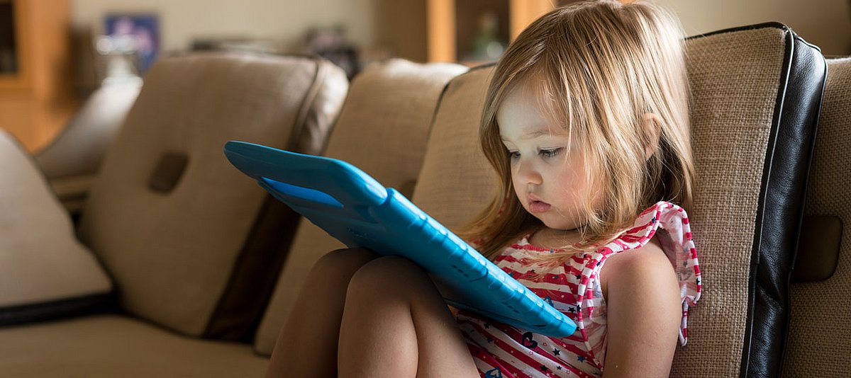 Ein kleines Mädchen sitzt zu Hause auf der Couch und schaut gebannt auf ein Tablet.