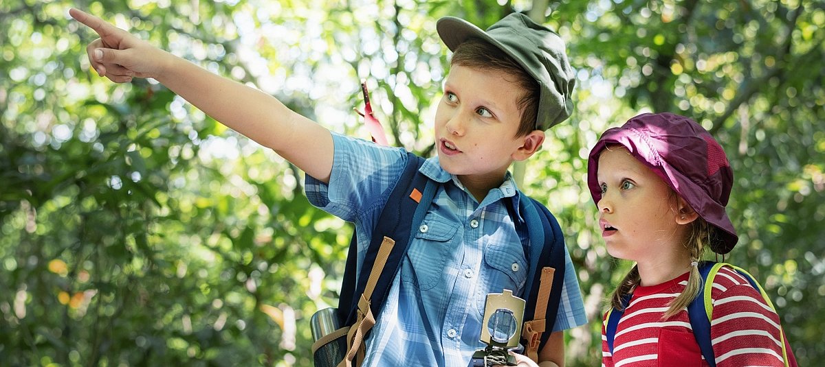 Ein Junge zeigt mit ausgestrecktem Arm auf eine Entdeckung in der Natur, das Mädchen neben ihm schaut gespannt