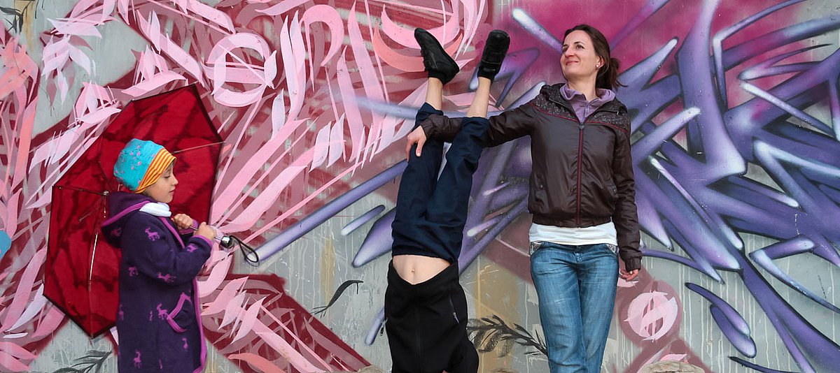 Eine Mutter steht mit ihren zwei Kindern vor einer Mauer mit einem Graffiti und hält die Beine ihres Kindes, das einen Handstand macht.  