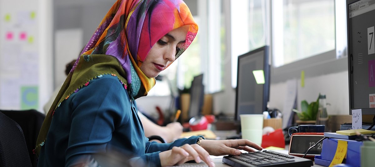 Junge Frau mit buntem Kopftuch sitzt an einem Schreibtisch und arbeitet am Computer