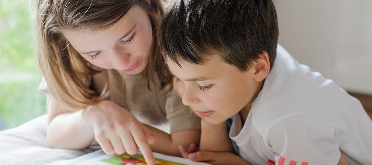 Zwei Kinder im unterschiedlichen Alter beugen sich über eine Buch und lesen.