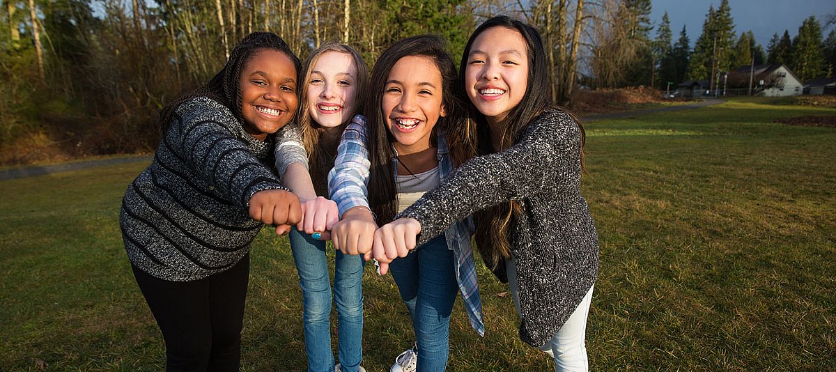 Vier junge Mädchen unterschiedlicher Herkunft stehen in einem Garten und stecken lächelnd die Fäuste zusammen.