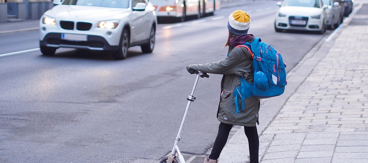 Mädchen steht mit City Roller am Straßenrand und möchte Straße überqueren