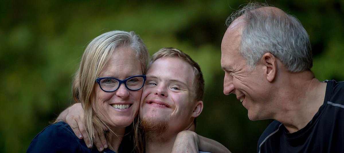 Auf dem Foto sind Eltern mit ihrem Sohn zu sehen, die sich in den Armen liegen und lachen. Der Sohn hat Down-Syndrom.