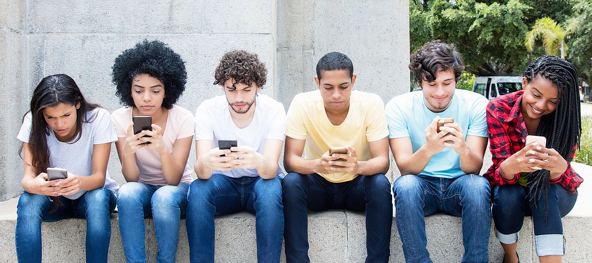 Junge Erwachsene unterschiedlicher Herkunft sitzen nebeneinander und schauen auf ihr Smartphone.