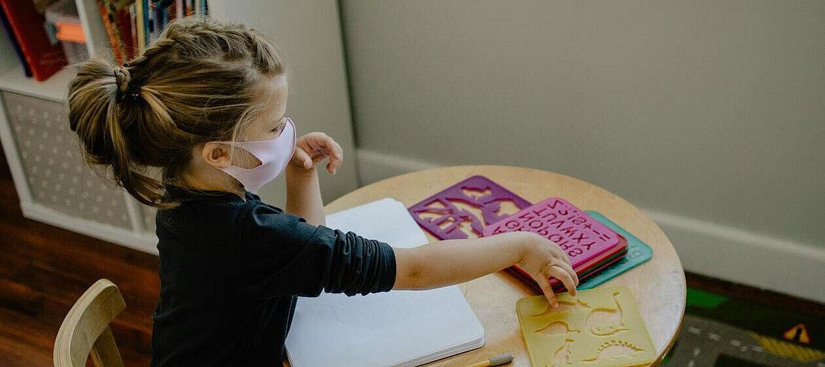 Ein Kindergartenmädchen übt Zahlen an einem Tisch. Sie trägt einen Mund-Nase-Schutz.