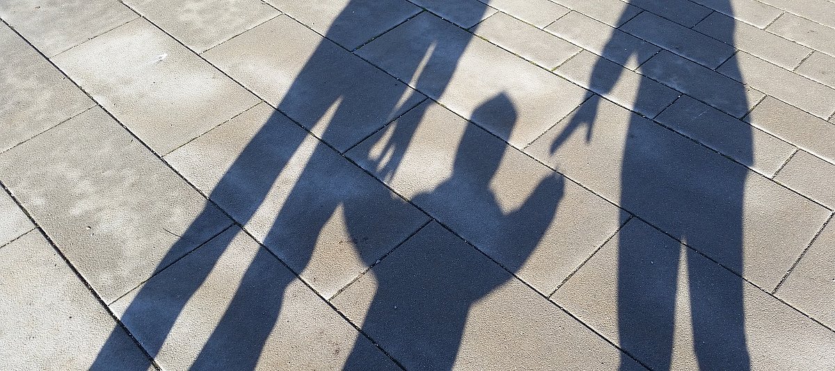 Die Schatten von zwei Erwachsenen, in deren Mitte ein Kind geht und nach der Hand des einen Erwachsenen greift