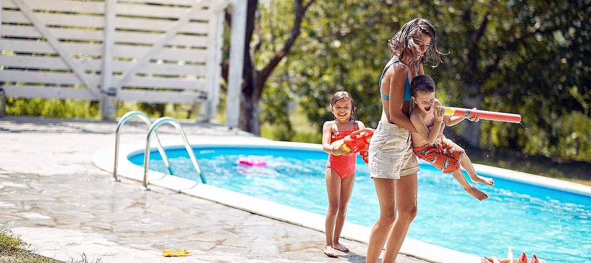 Eine Erwachsene spielt mit zwei Kindern am Rand eines Pools