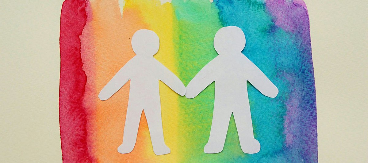 Zwei Papiermännchen auf einer Regenbogenfahne halten sich an den Händen.