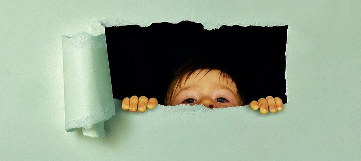 Ein Junge schaut mit der Nasenspitze durch ein in einer Papierwand aufgerissenes Fenster. 