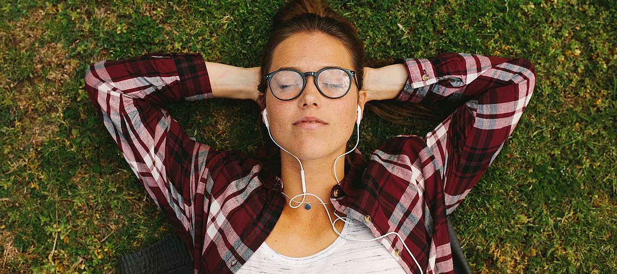 Eine jugendliche Person liegt mit geschlossenen Augen auf dem Gras und hat Kopfhörer in den Ohren