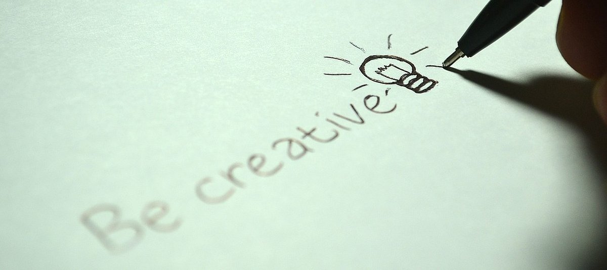 Eine Hand schreibt auf hellgrünen Untergrund "Be creative" und daneben ist eine Glühbirne gezeichnet